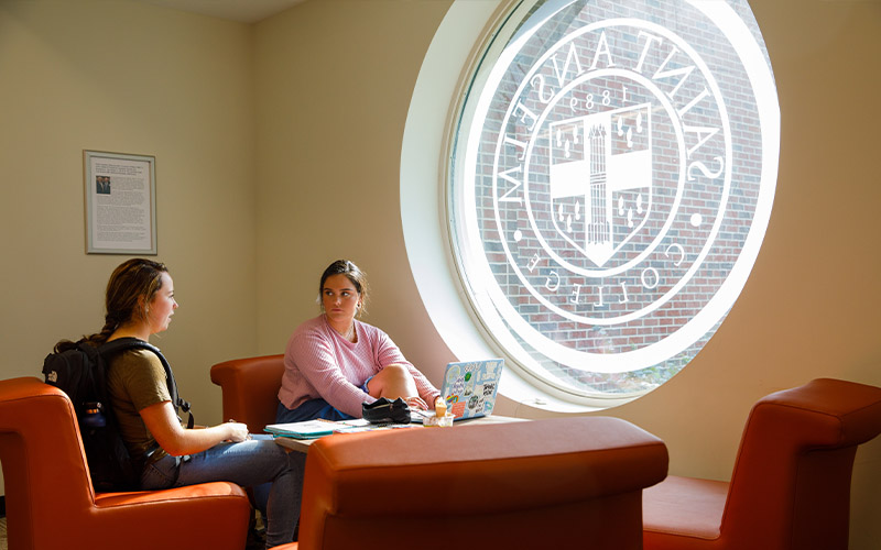 students sit in the Jean Student Center in front of a window with the Saint Anselm College seal
