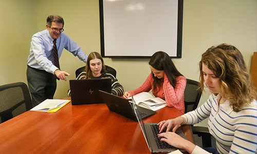 three students conduct research on their laptops with their professor at a table