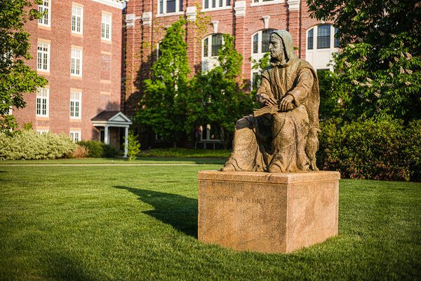 Stone statue of a man (Saint Benedict) in religious robes on a grassy lawn in front of brick buildings.