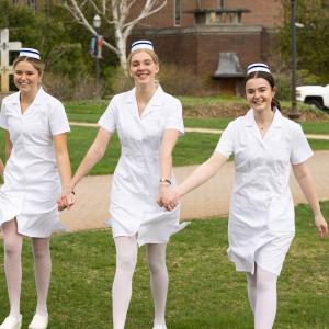 Nurses in pinning ceremony uniforms, holding hands while walking on Alumni Quad