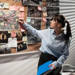 A women pointing to a picture on a corkboard with red threaded pins running in various directions