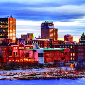 View of a lit mill city at dawn (or dusk) under a cloudy sky overlooking a rocky river.