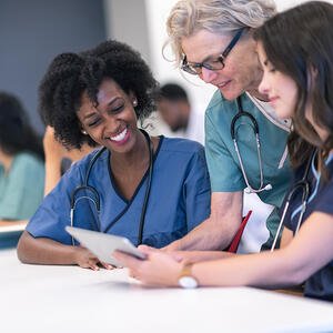 Group of female healthcare professionals in the foreground, collaborating around a tablet in a classroom setting.