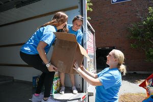 Three college women in light blue shirts smiling and unloading a box from a truck