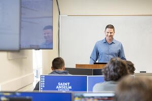 Male professor stands in front of a room of students and computers that show blue screens that say "Saint Anselm College" on them