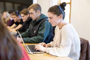 Young woman with brunette hair sitting in front of a laptop in a room with other students
