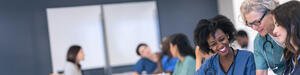 Group of female healthcare professionals in the foreground, collaborating around a tablet in a classroom setting.